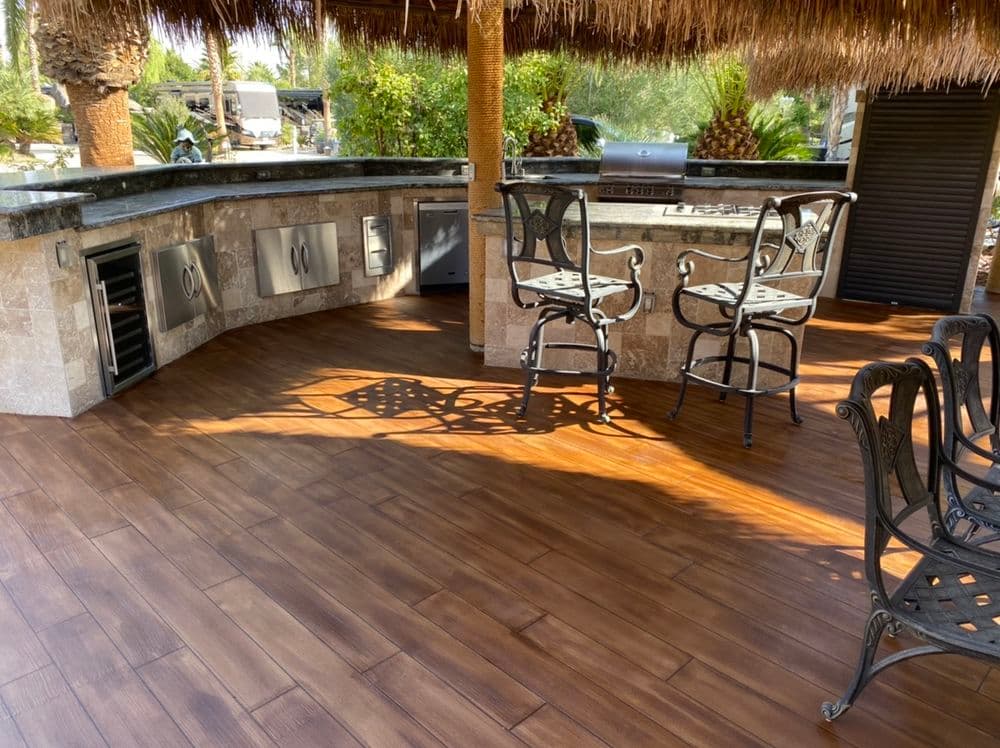 Outdoor kitchen with bar stools, modern appliances, and wooden tiles under a thatched roof.
