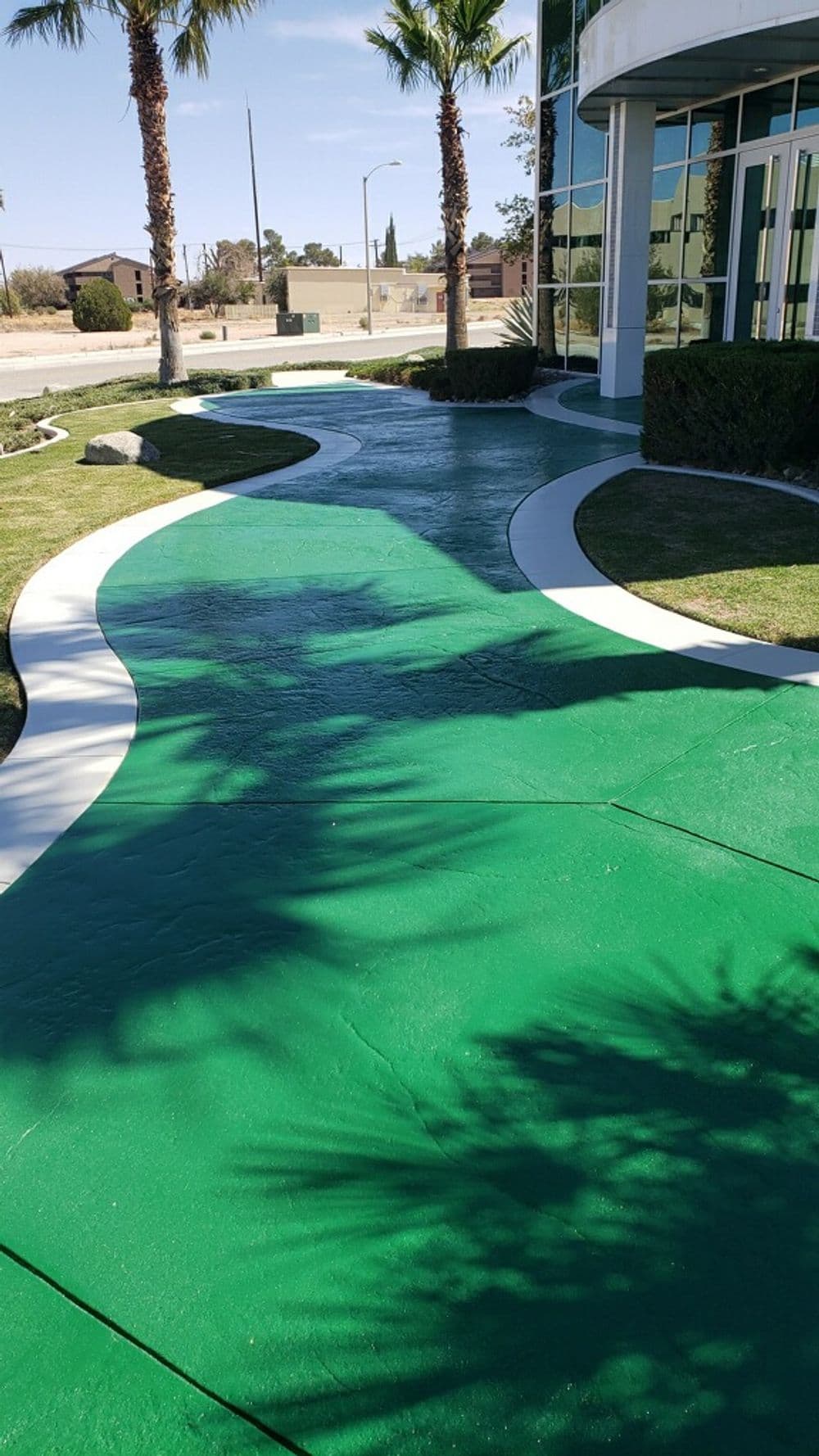 Green concrete pathway surrounded by palm trees leading to a modern building exterior.
