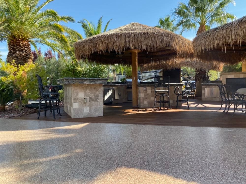 Tropical outdoor kitchen with thatched roof, stone bar, and palm trees in a sunny setting.