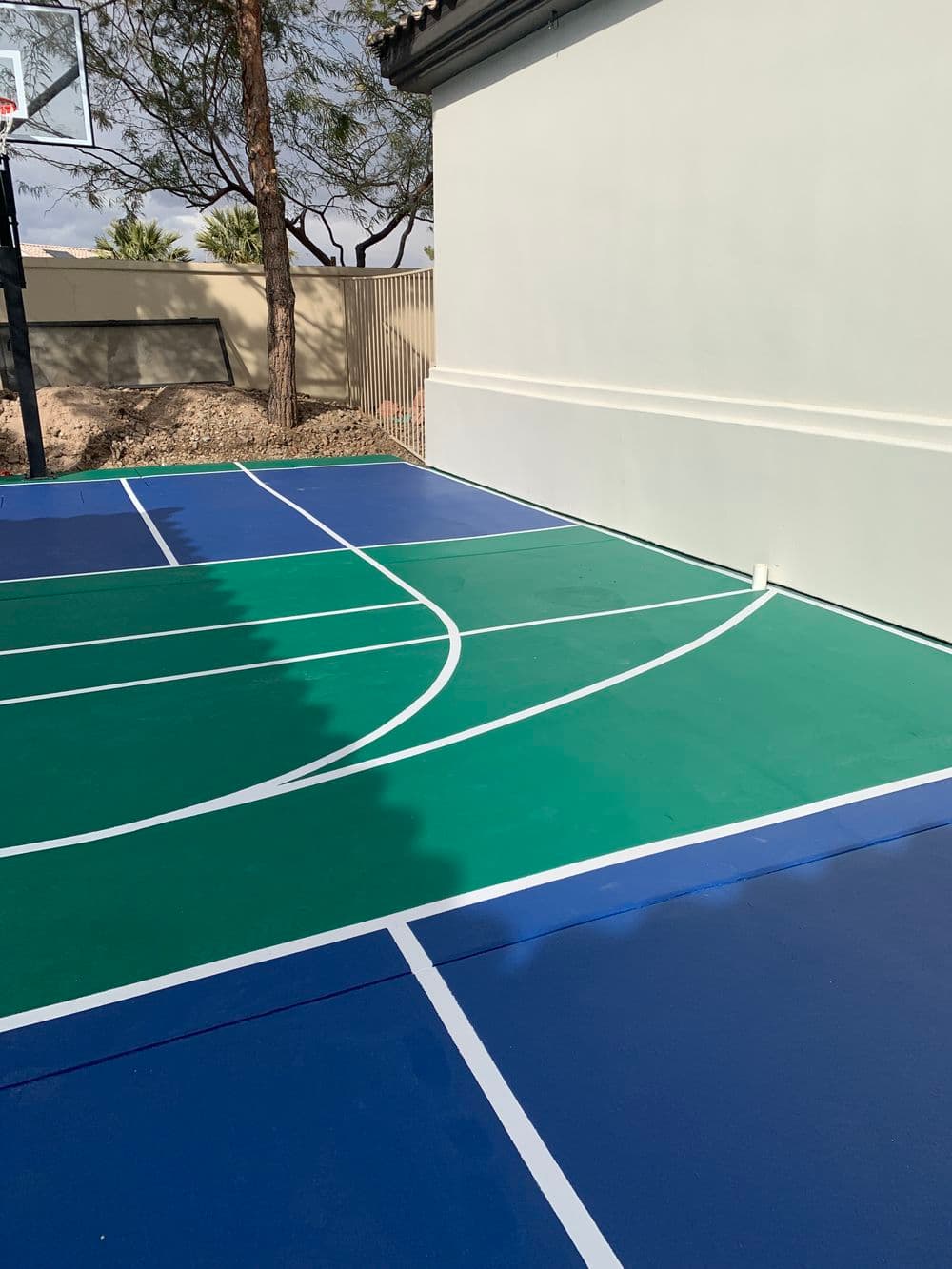Colorful outdoor basketball court with green and blue sections beside a wall and trees.